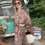 Woman in a patterned robe standing in front of a vintage car with a colorful rug and pillows on the ground.