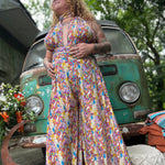 Person wearing a colorful floral one piece outfit standing in front of a vintage van with greenery around.