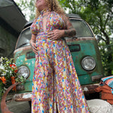 Person wearing a colorful floral one piece outfit standing in front of a vintage van with greenery around.