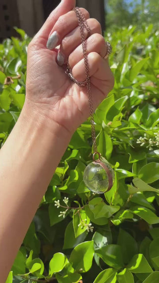 Someone holding the Crystal Ball Necklace in Copper - Clear Quartz in front of greenery.