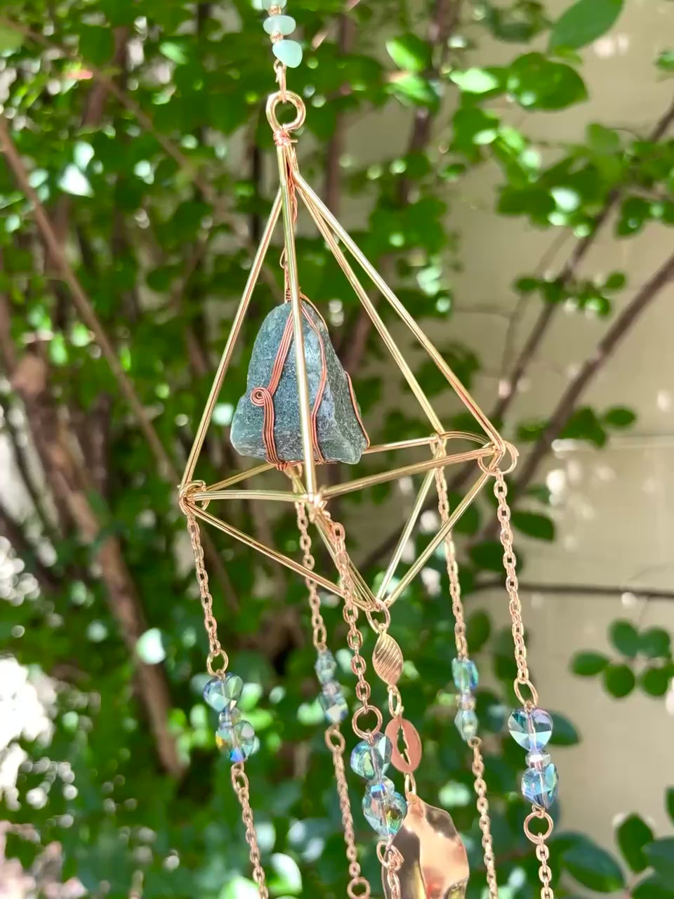 Someone holding the Amazonite Lotus Crystal Suncatcher in front of a cream-colored brick wall with greenery.