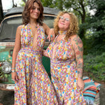 Two women in colorful floral jumpsuits standing next to a vintage car with a green leafy background.