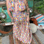 Person wearing a colorful patterned jumpsuit in front of a vintage vehicle with a relaxed outdoor setting.