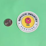 The Service Industry Restaurant Humor Sticker next to a quarter on a green background.