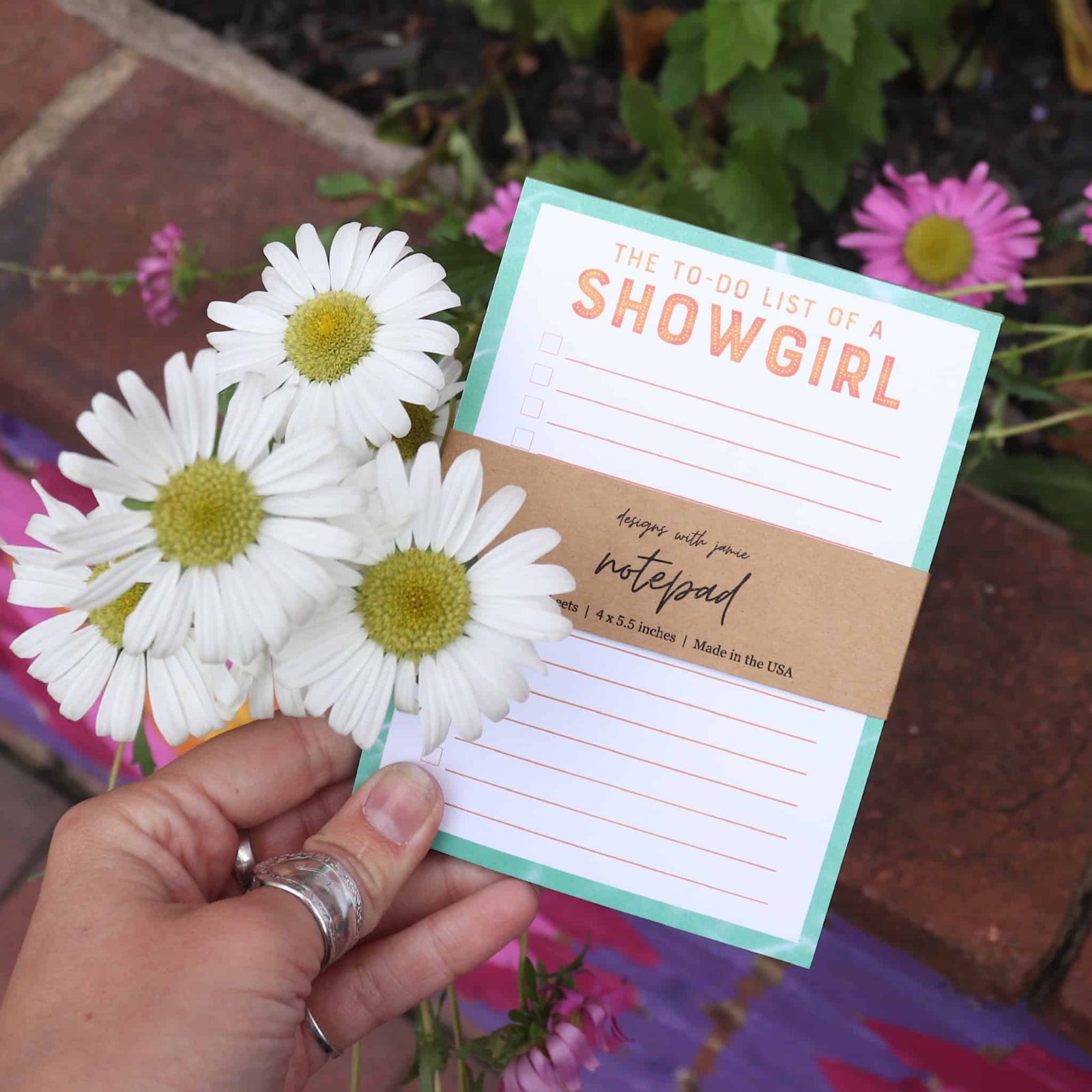 Hand holding a notepad with 'The To-Do List of a Showgirl' text, surrounded by white flowers.