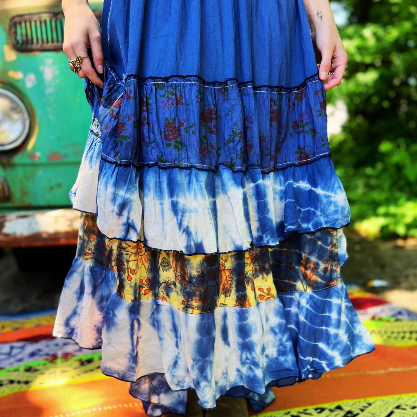 Person wearing a colorful, tiered dress with a tie-dye pattern in front of a green vehicle.