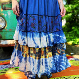 Person wearing a colorful, tiered dress with a tie-dye pattern in front of a green vehicle.