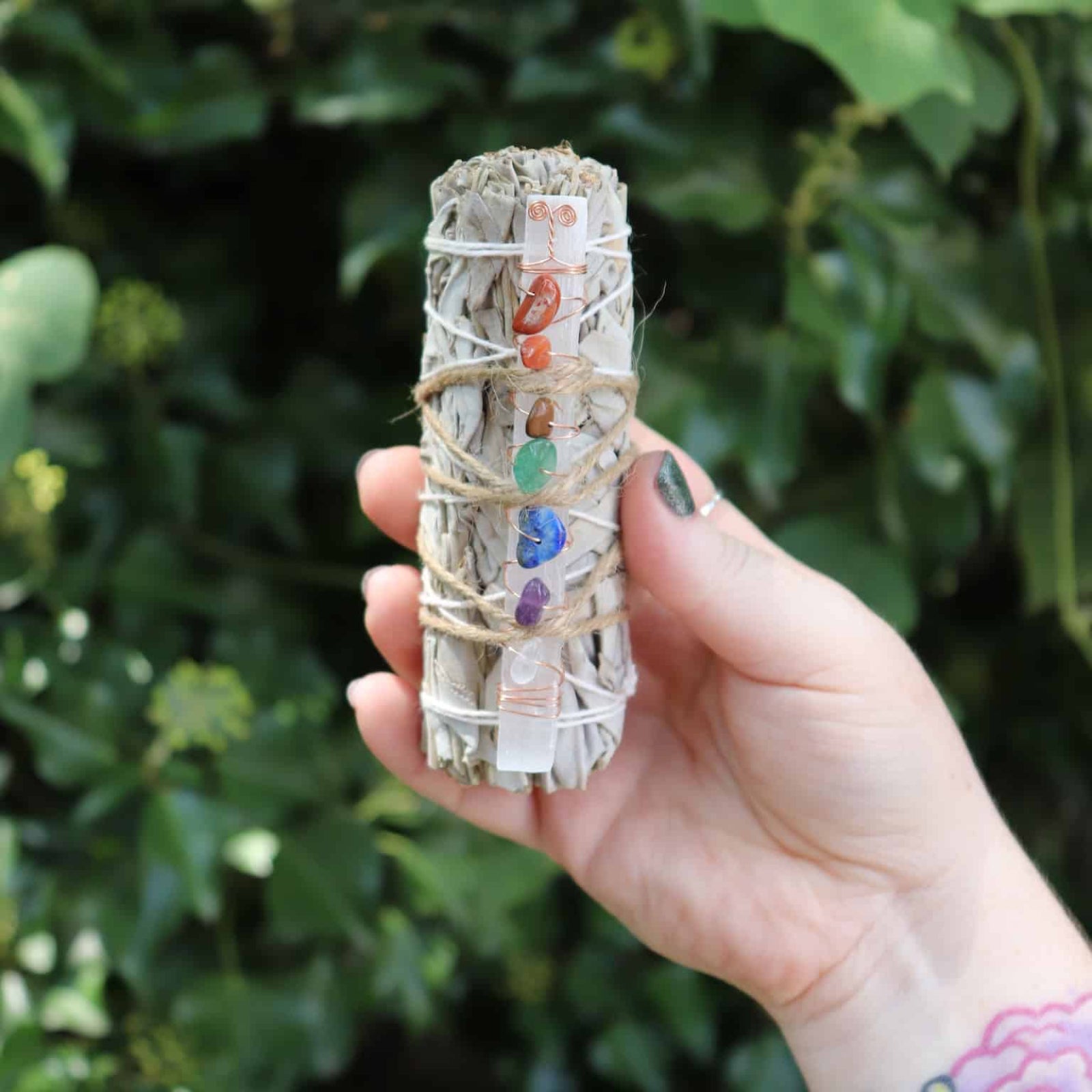 Hand holding a smudge stick with colorful stones against a green leafy background