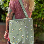 Blonde woman wearing a floral dress and carrying a sage green linen tote bag with hand-embroidered white daisies
