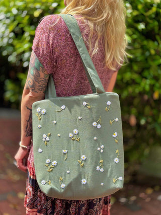 Blonde woman wearing a floral dress and carrying a sage green linen tote bag with hand-embroidered white daisies